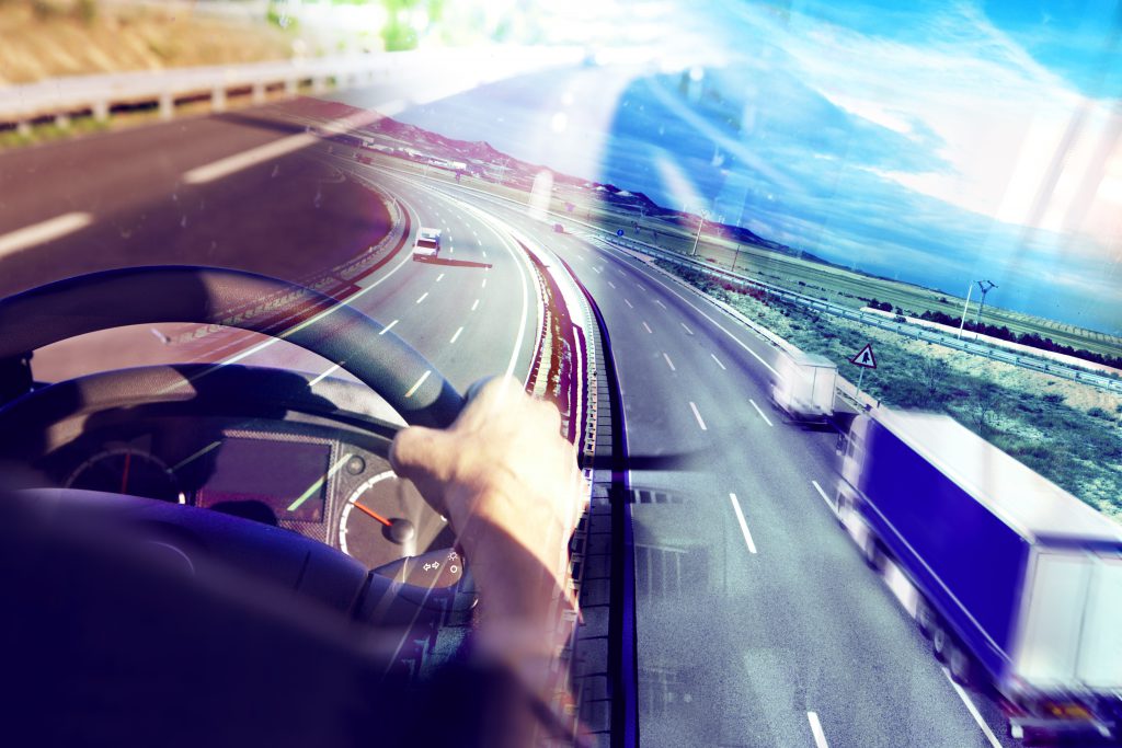 Hands holding onto a steering wheel of a large truck looking out on the road.